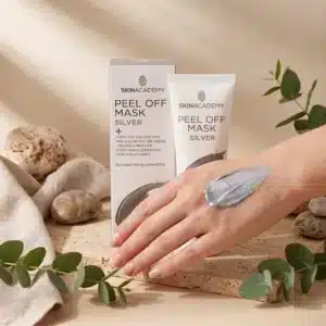 Iholle sivelty metallinhohtoinen hopeinen Skin Academy Silver Peel Off Mask -kasvonaamio kädenselässä. Taustalla tuotteen pahvipakkaus, valkoinen tuubi, eukalyptuksen oksia ja luonnonkiviä.
