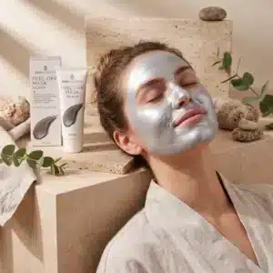 Nainen makaa silmät suljettuina hopeinen kasvonaamio kasvoillaan spa-ympäristössä. Taustalla on Skin Academy Silver Peel Off Mask 80 ml -tuotepakkaus, tuubi, eukalyptuksen oksia ja luonnonkiviä.