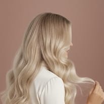 Naisen pitkät, vaaleat ja laineikkaat hiukset takaapäin kuvattuna vaaleanpunaisella taustalla.