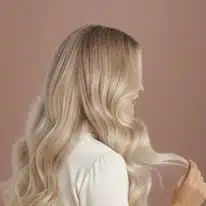 Naisen pitkät, vaaleat ja laineikkaat hiukset takaapäin kuvattuna vaaleanpunaisella taustalla.