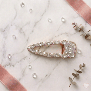 Kimalteleva kullanvärinen hiusklipsi, jossa on runsaasti kirkkaita ja sateenkaariväreissä hohtavia strassikiviä, aseteltuna valkoiselle marmoripinnalle.