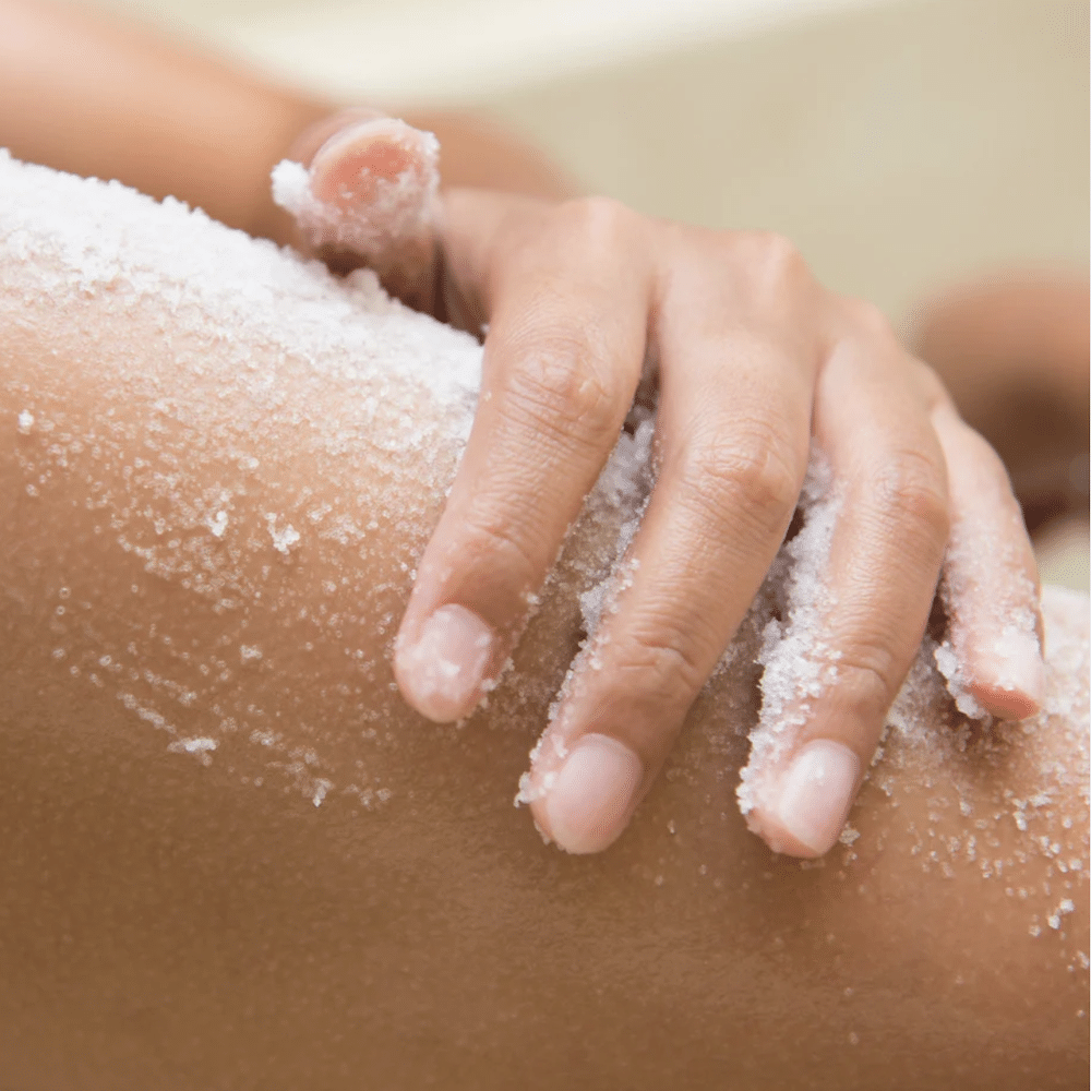 Lähikuva kädestä, joka levittää karkeajakoista valkoista vartalonkuorinta-ainetta säären iholle. Kuorintasuolan kiteet näkyvät selvästi kostealla ja pehmeällä iholla.