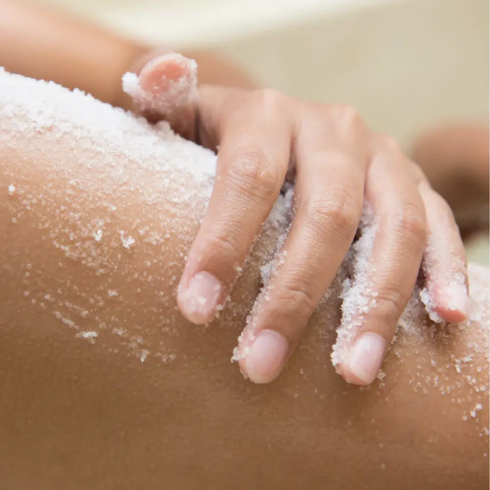 Lähikuva kädestä, joka levittää karkeajakoista valkoista vartalonkuorinta-ainetta säären iholle. Kuorintasuolan kiteet näkyvät selvästi kostealla ja pehmeällä iholla.