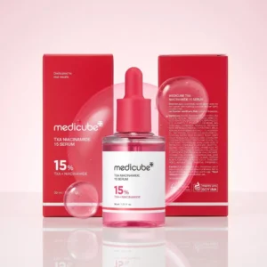 Medicube TXA Niacinamide 15 Serum -lasipullo asetettuna keskelle kiiltävää tasoa, taustallaan kaksi punaista pakkauslaatikkoa etu- ja takapuolelta kuvattuna, suurten läpikuultavien nestekuplien ympäröimänä.