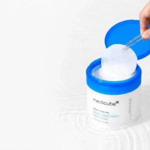 Avattu sähkönsininen Medicube Zero Pore Pad -purkki asetettuna kevyesti väreilevään veteen, josta käsi nostaa läpinäkyvillä pinseteillä pyöreää, kohokuvioitua lappua kirkkaan valkoisella taustalla.